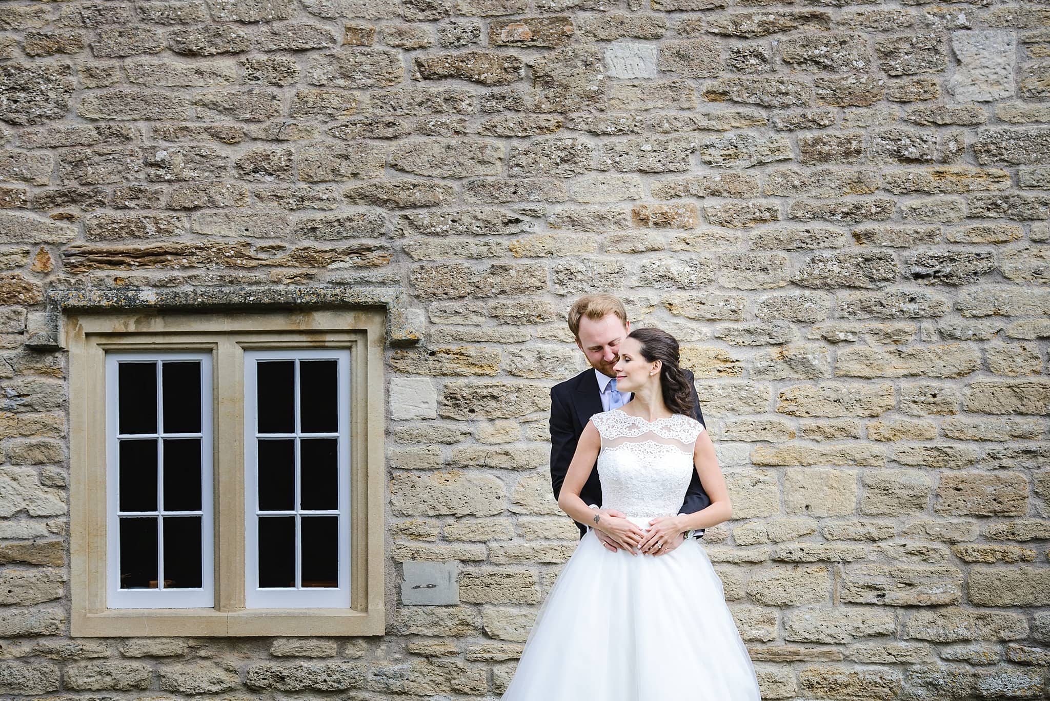 Bride and groom embrace each other in front of a brick wall and window outside Wick Farm