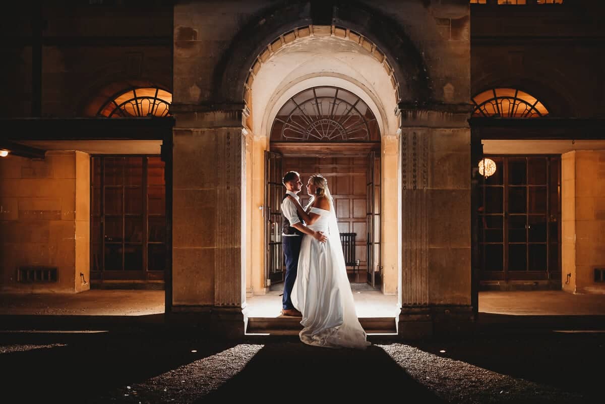Bride and groom stand in archway of Coombe lodge at night back lit