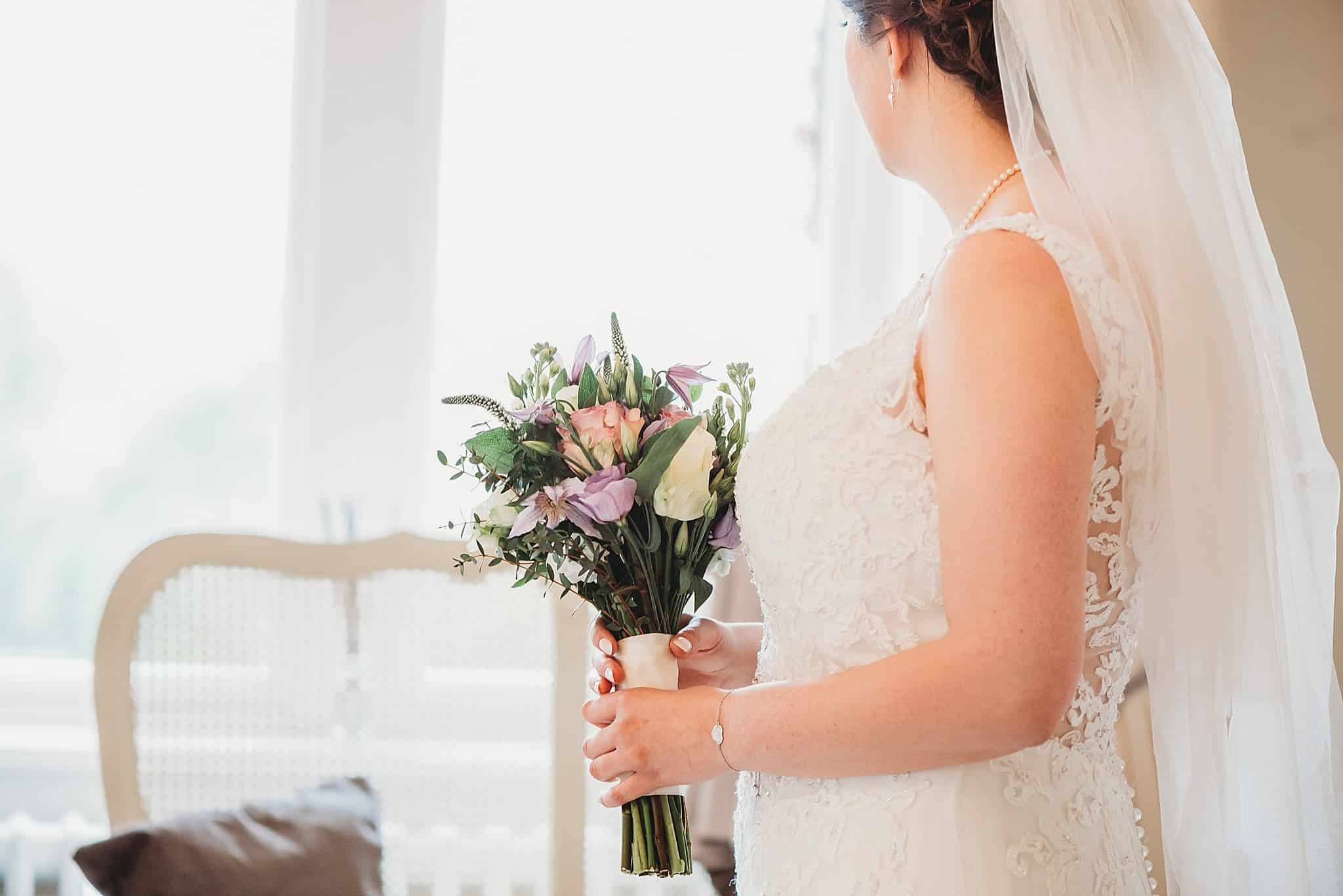 bride holding her bouquet of flowers looking out of the window in the bridal suite at Manor by the lake wedding venue