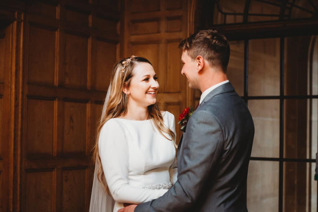 Bride smiles at Groom inside Coombe Lodge with brown wooden panels in the background.