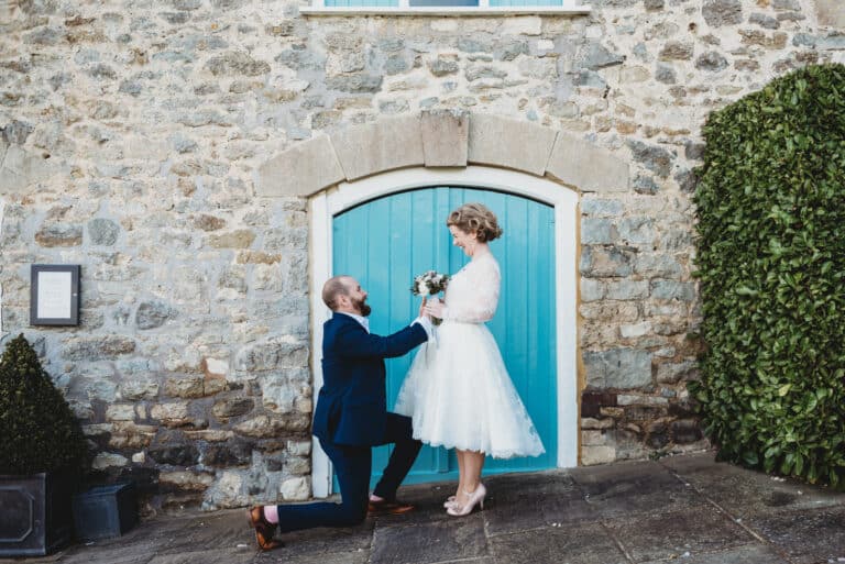 groom goes on one see presenting to bride in front of the blue door